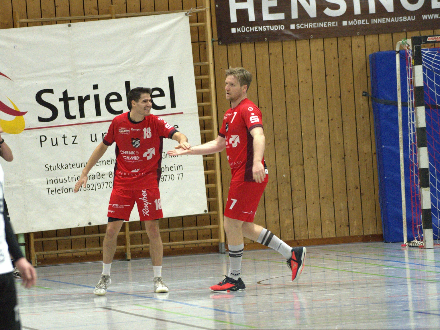 Zwei Handballspieler des HRW klatschen sich freudig in einer Turnhalle ab. Im Hintergrund sind ein Torpfosten und Sponsorenplakate zu sehen. Auf der linken Seite ist eine Person angeschnitten.