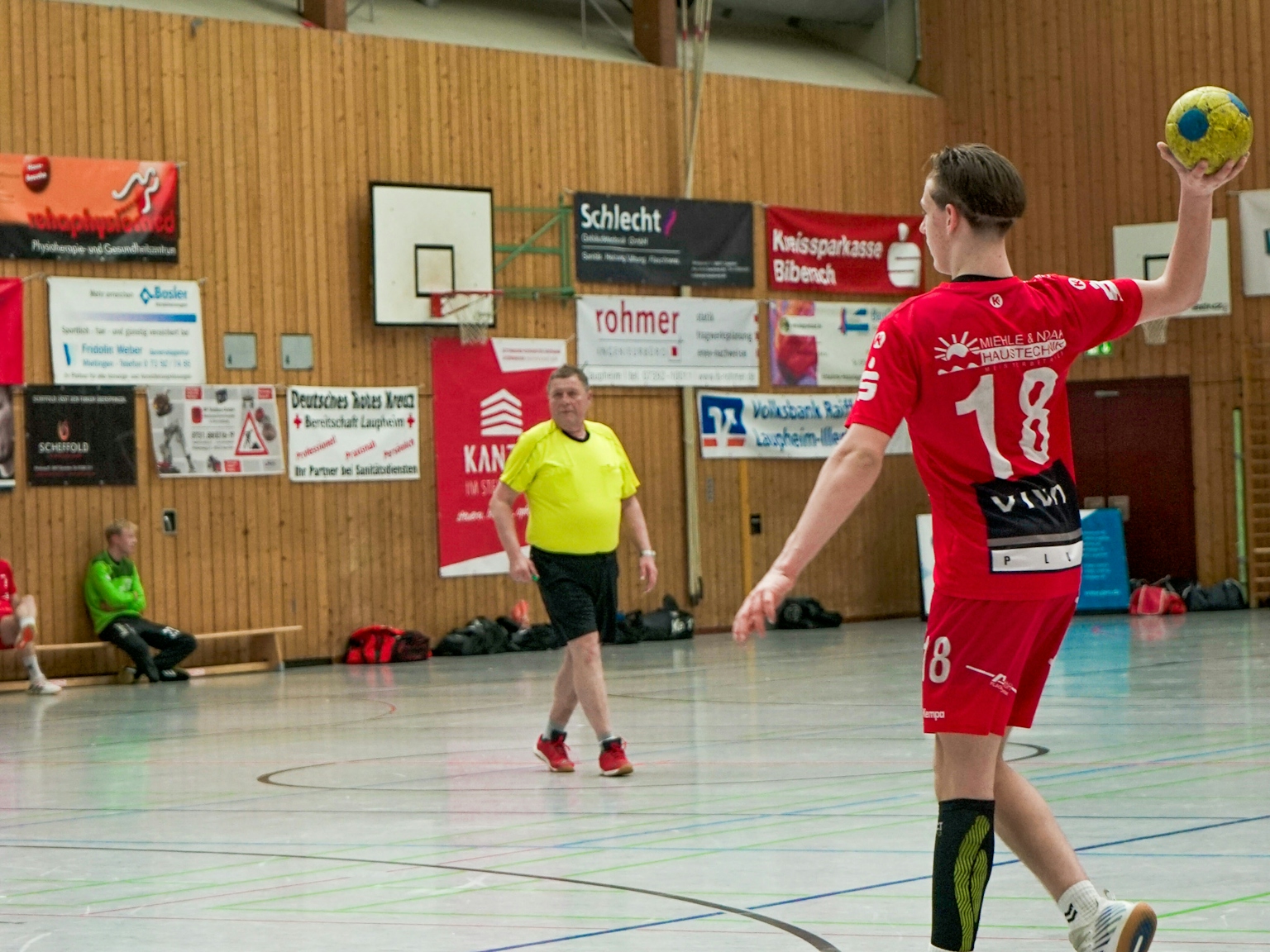 In einer Turnhalle,im Vordergrund auf der rechten Seite ist ein Handballspieler des HRW mit rotem Trikot im Begriff den Ball zu werfen. im Mittelgrung im Zentrum des Bilds steht der Schiedsrichter mit gelbem T-shirt. Im Hintegrund sind Auswechselspieler auf einer Bank und Sponsorenplakate zu sehen.