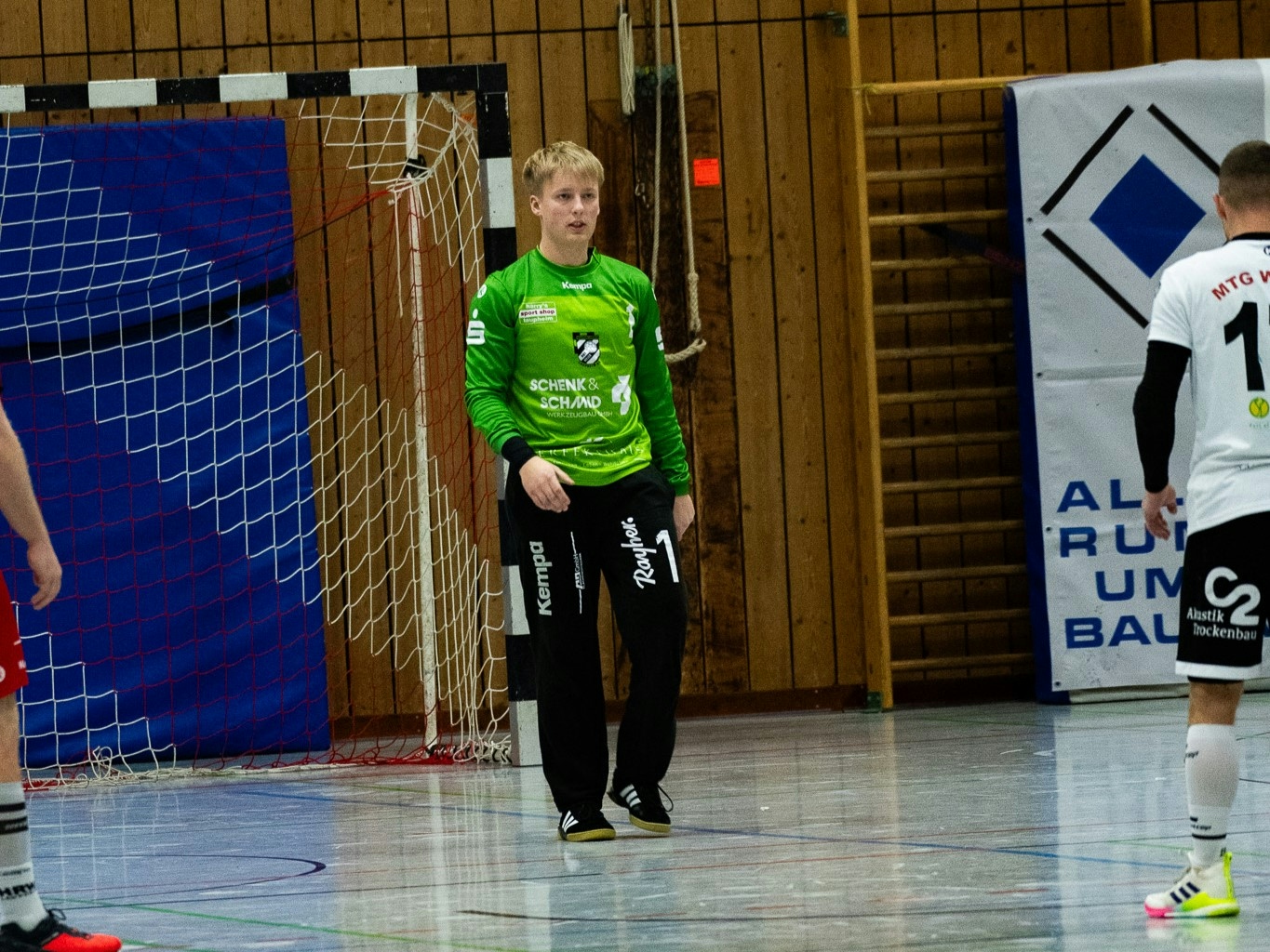 Handball Torwart mit grünem Trikot im Mittelpunkt in einer Sporthalle. Im Hintergrund ist das Tor zu sehen, am linken und rechten Rand des Bildes steht jeweils ein weiterer Spieler